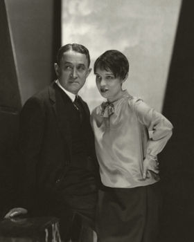 A man in a suit and a woman in a satin blouse stand closely in a dimly lit room, conveying a serious mood. Black and white tones dominate.