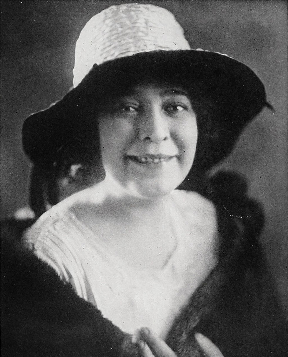Smiling woman in a wide-brimmed hat and fur stole, black and white setting, evokes a vintage, elegant mood. Background is blurred.