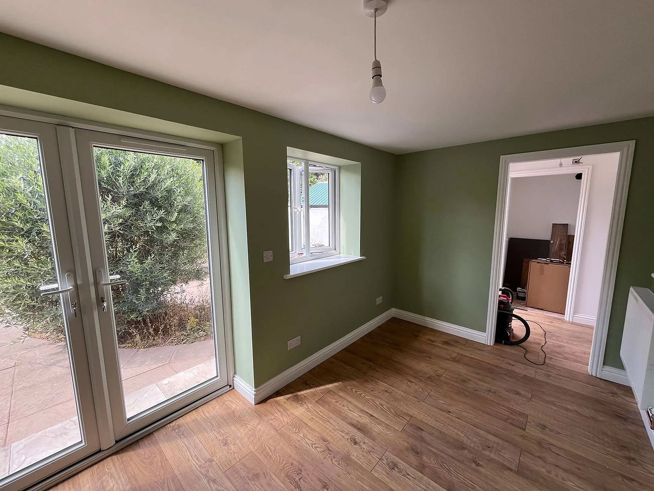 A room with light green walls and wooden flooring, featuring large glass doors opening to a garden