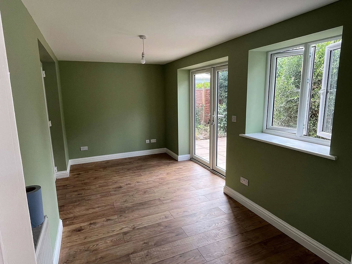 A bright, empty room with green walls and wood flooring features a large window and glass door