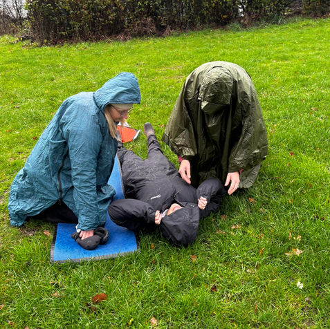 Two people in raincoats assist a person lying on the grass