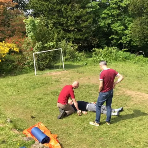 A man in a red shirt aids a person lying on the grass in a bright