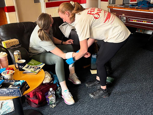 A woman assists another sitting on a sofa, helping her with arm exercises
