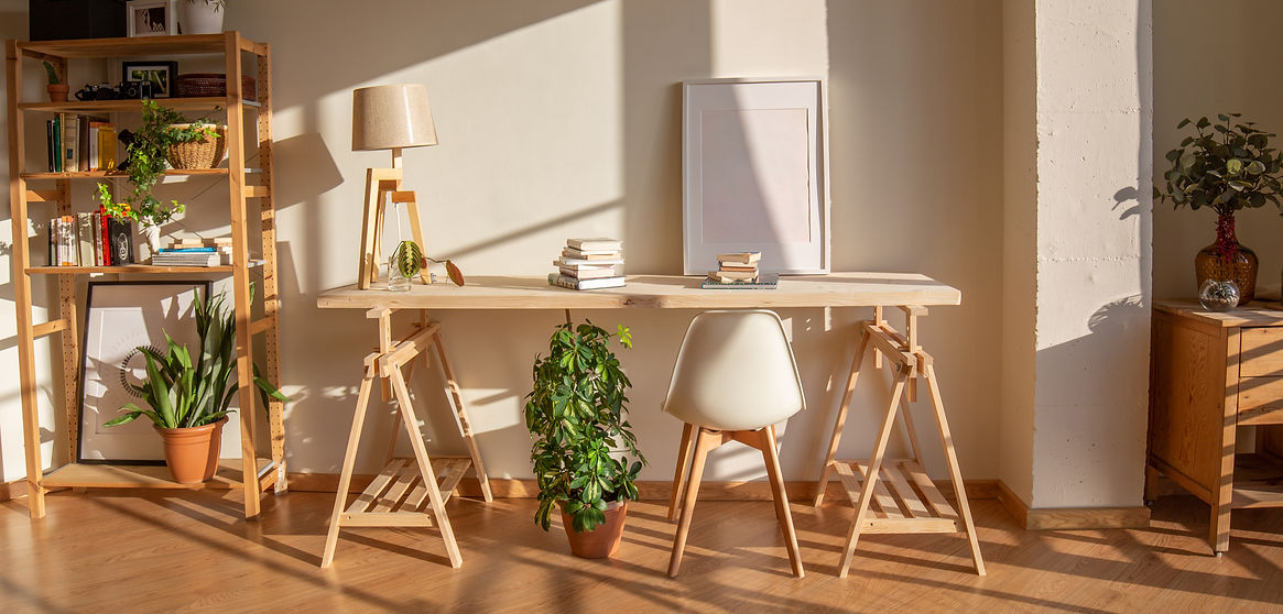 Sunlit minimalist home office with a wooden desk, a white chair, stacked books, a potted plant, bookshelves