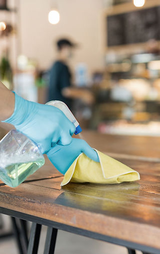 A person wearing blue gloves cleans a wooden table with a spray bottle and yellow cloth