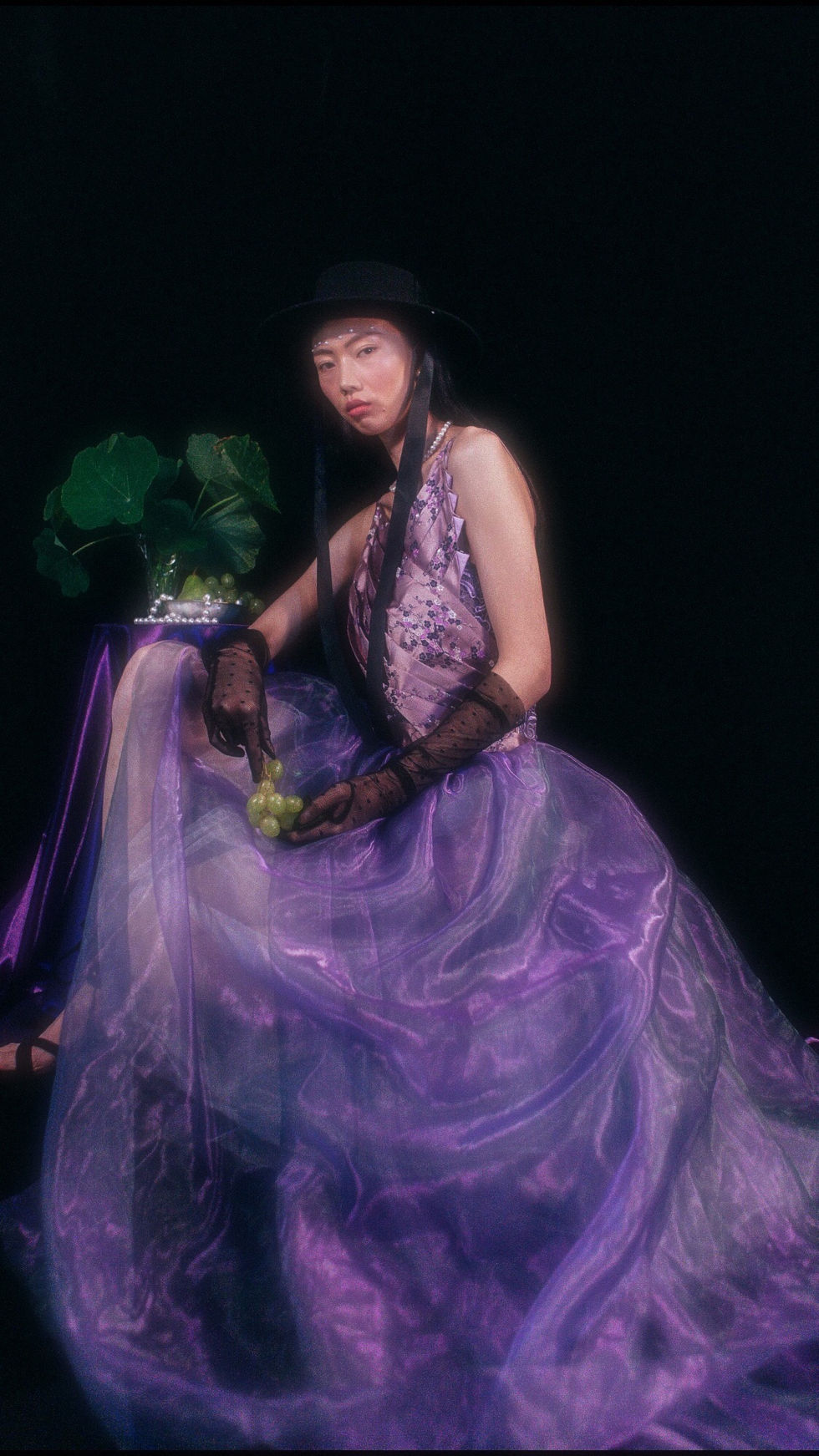 Woman in purple gown and black hat holding grapes.