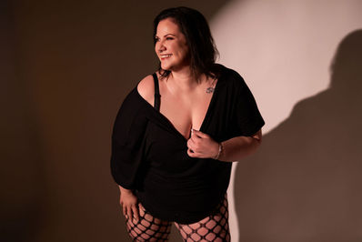 Smiling woman in black top and fishnet tights poses in a studio, lit by warm light with her shadow on the wall.