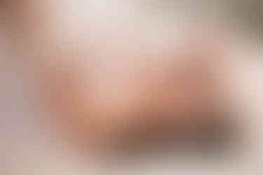 Boudoir photo of woman laying on bed covered by white sheets. Her eyes are closed and one hand in her hair.