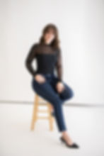 Woman in black top and jeans sits on a wooden stool, smiling against a plain white background. The Studio Manager for Boudoir Photography Studio in the Minneapolis Metro.