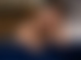 Woman with wavy hair leans on plush blue sofa, smiling warmly. Soft, blurred background with lights adds cozy ambiance.