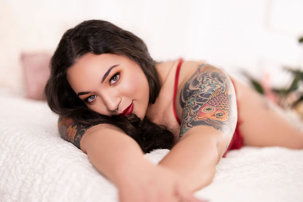 Woman in red lingerie with tattoos lies on a white bed, smiling softly. Bright tattoo on arm, blurred cozy room background.