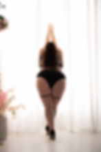 A woman in black lingerie and heels stands in front of sheer curtains, reaching up. Floral arrangements on either side add softness.