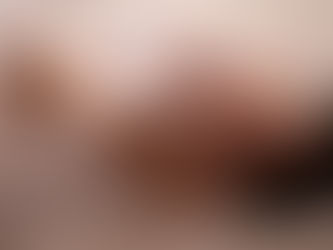 Woman lying on a bed under white sheets, finger near her lips, eyes closed, in soft warm light.