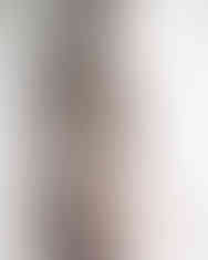 Bride in lace lingerie and veil stands near a bright window, gazing outside. Soft lighting and white tones create an intimate mood.