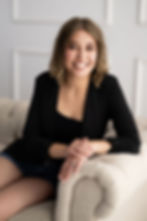 Smiling woman sitting on a beige couch, wearing a black outfit. She looks relaxed in a softly lit room with white paneled walls. MUA at Minneapolis Boudoir Studio.