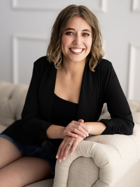 A woman in a black blazer and jean shorts poses on a cream colored couch. She is smiling and looks confident. 