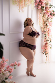 Woman stands next to white wall with flower arch over her in boudoir photography studio. She's wearing dark purple lingerie and her head is raised in confidence.