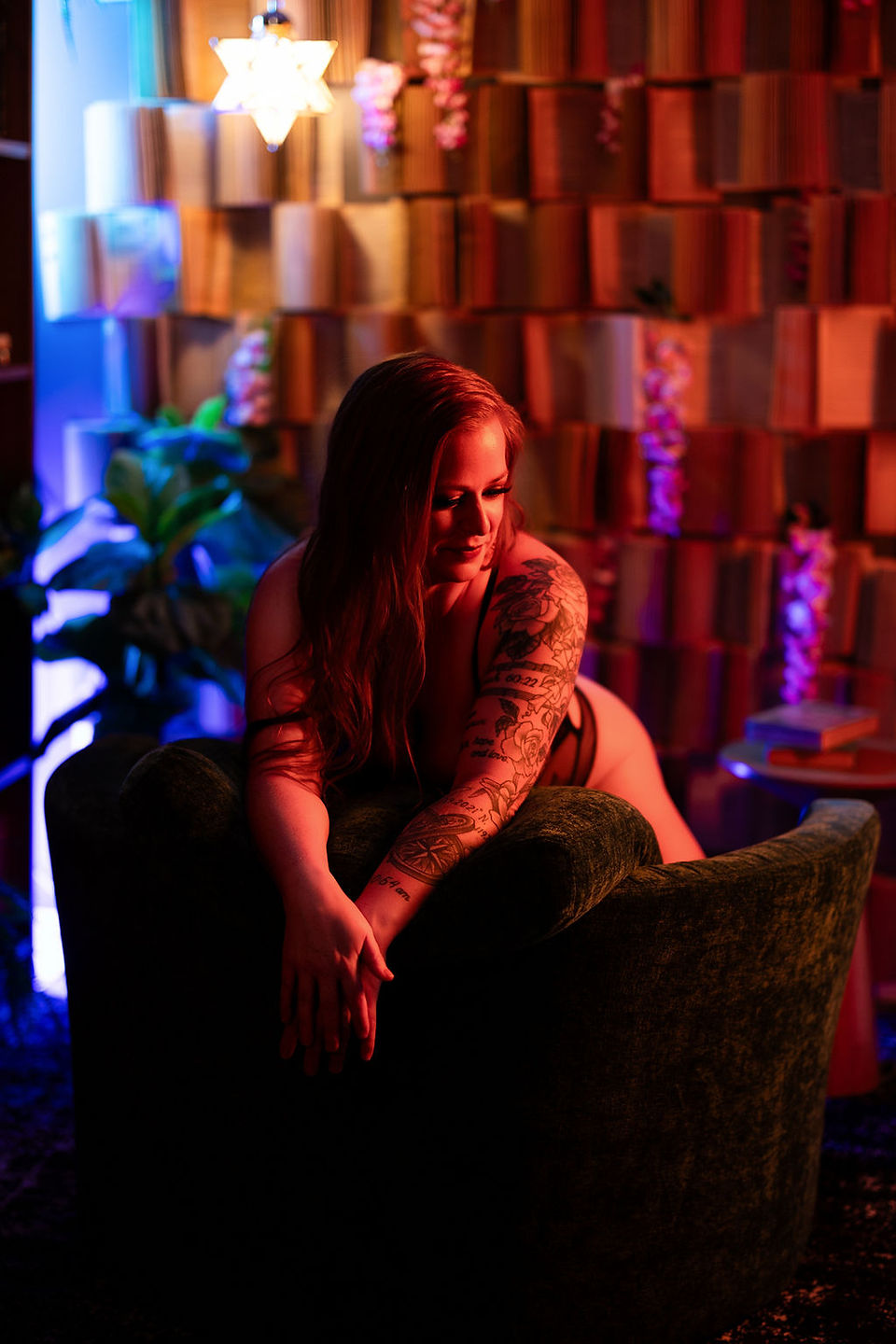 Tattooed woman reclining on a dark chair in a neon-lit lounge, with warm red and blue lighting and a patterned wall behind her