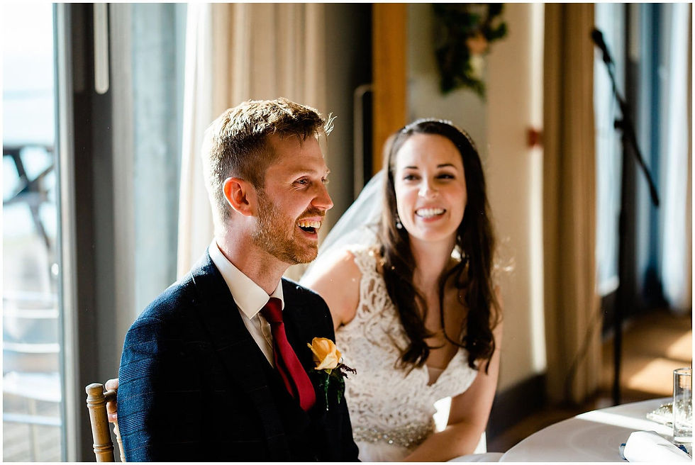 Smiling bride and groom at a sunlit indoor wedding reception, seated by a window with warm, joyful mood.