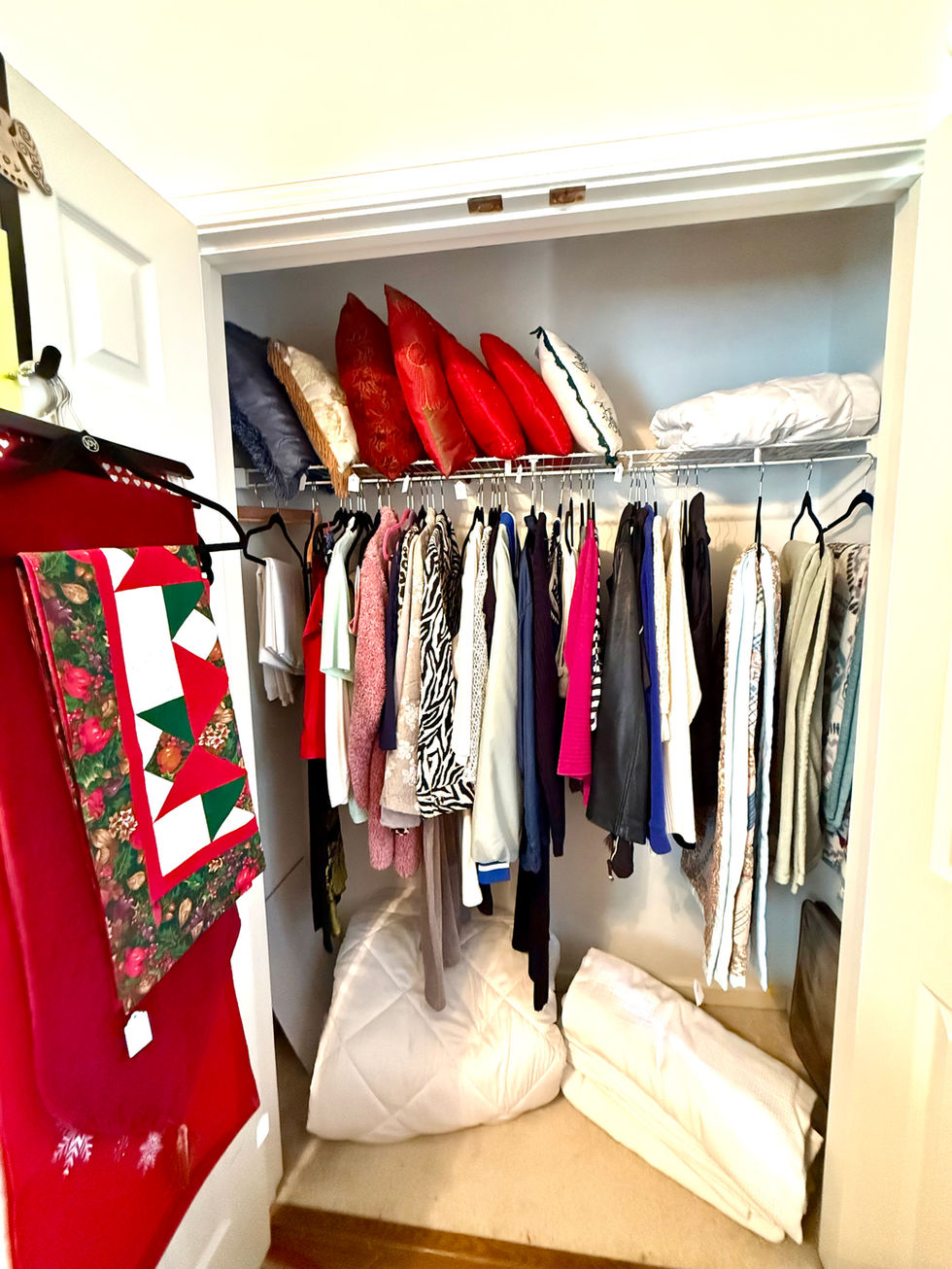 Closet with clothes, pillows, and a red quilt.