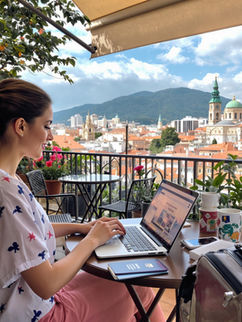A young professional working on a laptop at a cozy outdoor café overlooking a vibrant cityscape, with a suitcase and passport visible.