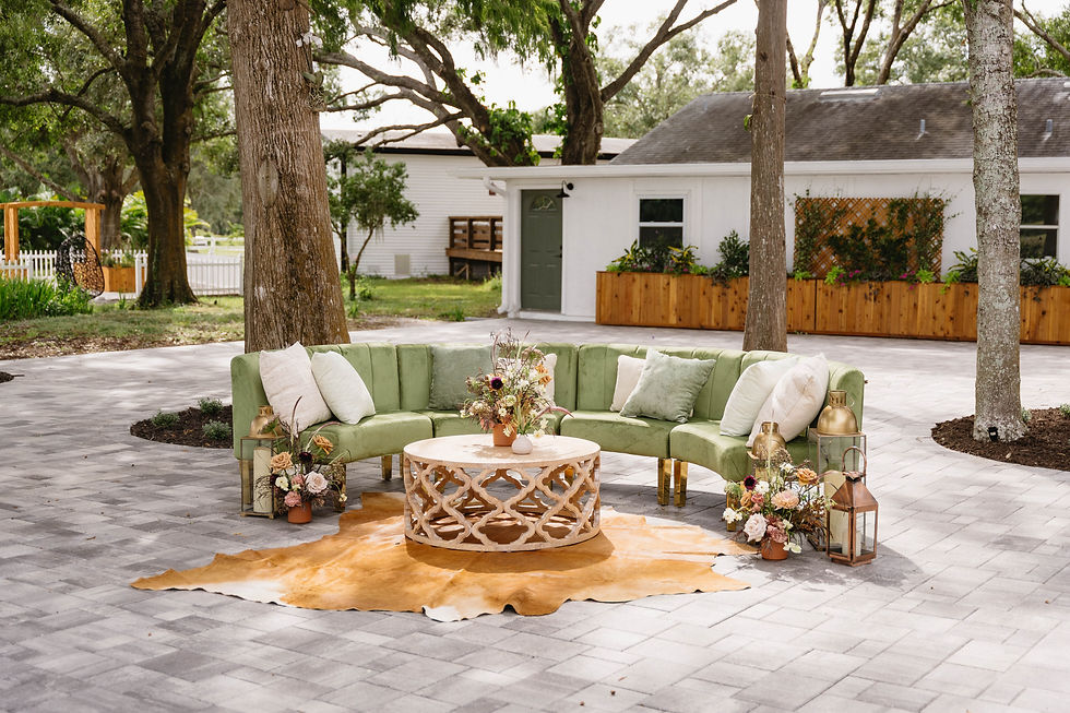 Outdoor lounge setup with green curved sofa, floral centerpieces, lanterns, and tan rug in a tree-shaded patio by a white house.