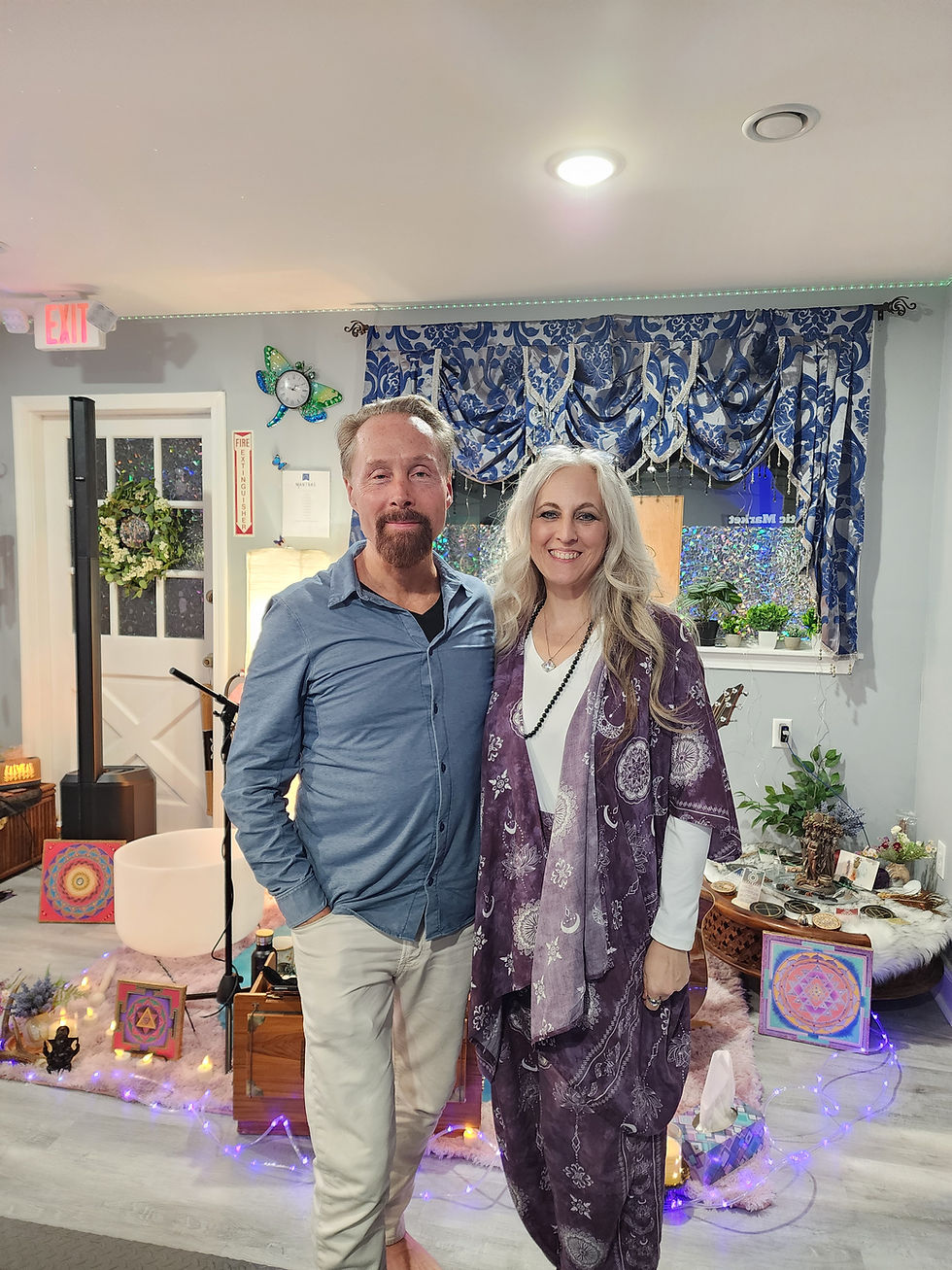 Girish and Laurie Gouley stand smiling in a room with mystical decor, including mandalas and fairy lights. Blue patterned curtains hang in the background.