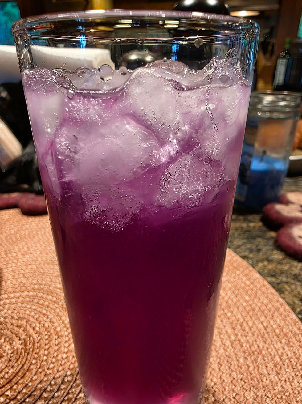 Tall glass of purple iced drink on a woven coaster in a kitchen, with ice cubes and a calm, casual feel.