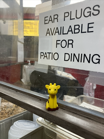 Giraffe toy sitting under a sign that says "EAR PLUGS AVAILABLE FOR PATIO DINING"