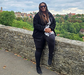 A person in black attire, wearing sunglasses, smiles while sitting on a stone wall. Green trees and colorful buildings fill the scenic background.