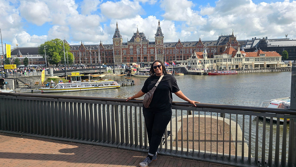 Smiling person in sunglasses leans on a railing by a canal. Historic building and boats in the background, with blue skies and clouds.