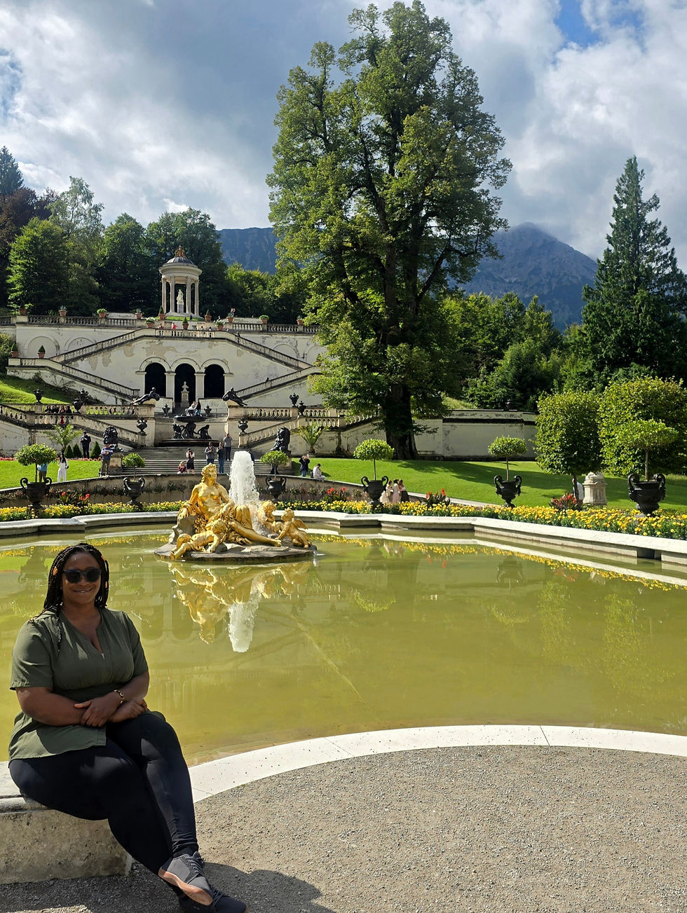 A woman sits by a fountain with golden statues in a lush garden, backed by a grand white staircase and a tree. Mountains are visible behind.