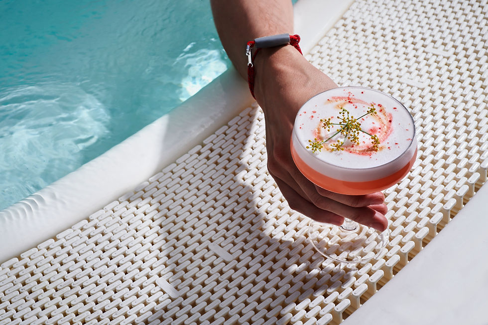 Hand holding a pink cocktail with foam and herbs beside a turquoise pool on a white woven floor. Sun Club, Virgin Voyages.
