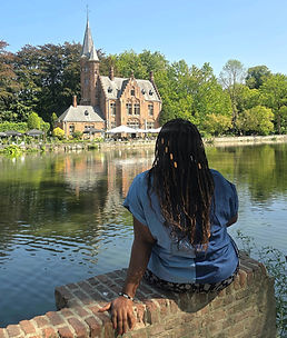 Woman in blue shirt sits by a lakeside brick wall, facing a castle-like building under a clear sky. The scene is serene and picturesque.
