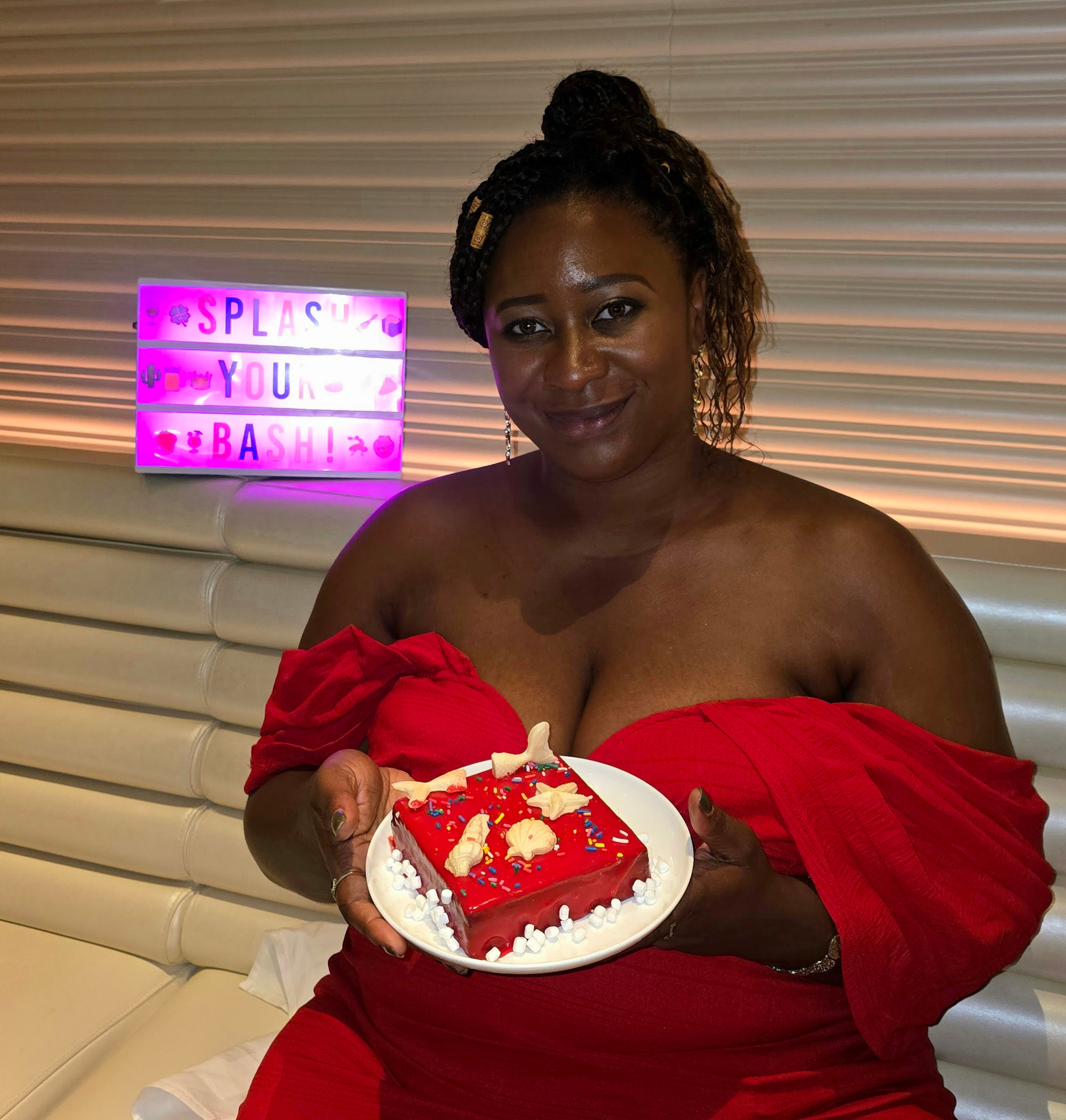 Virgin Voyages-The Wake-Smiling woman in red dress holds a decorated red dessert plate at a party, with a neon Splash Your Bash sign behind her.