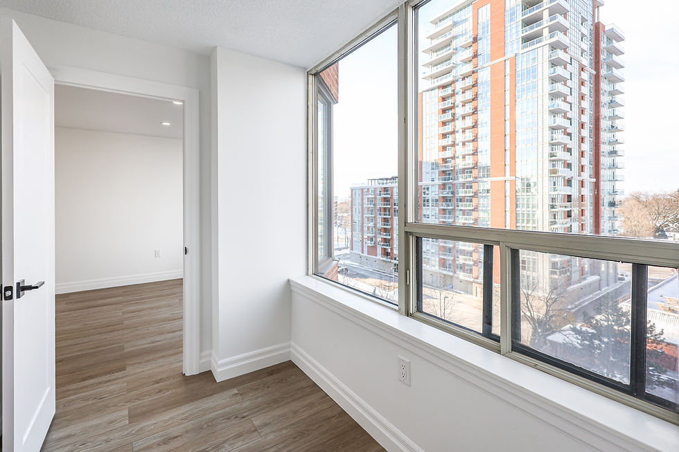 Bright room with wooden floor, large windows, and view of modern high-rise buildings. White walls and open door create a spacious feel.