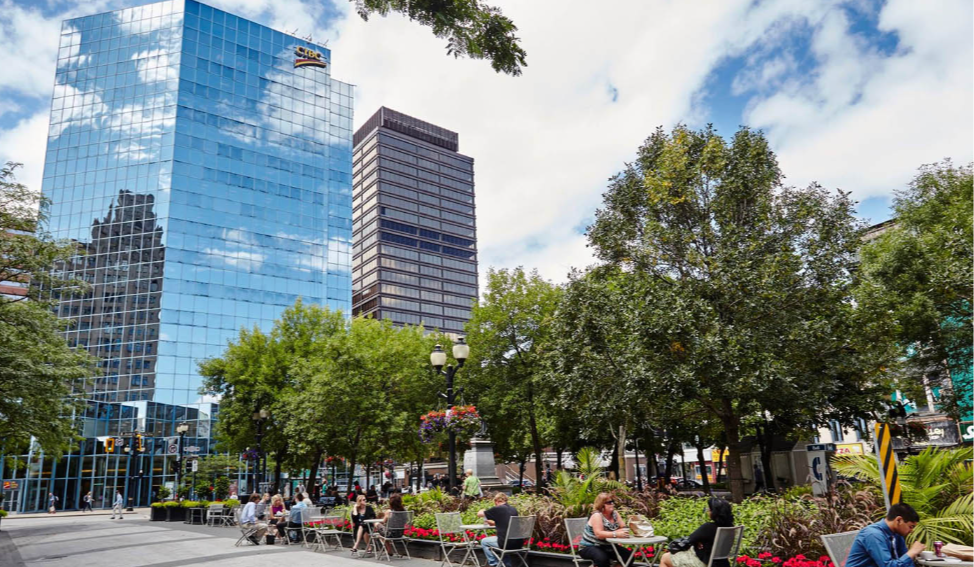 People sit at tables in a city plaza with trees and flowers. Reflective glass skyscraper and cloudy sky in background. Relaxed atmosphere.