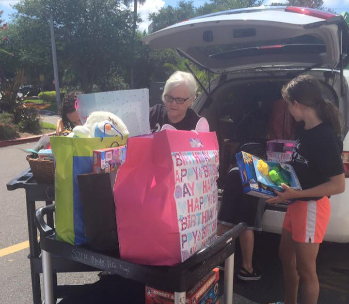 Two people load birthday gift bags and toys into a car. One bag reads "HAPPY BIRTHDAY." It's sunny with trees in the background.