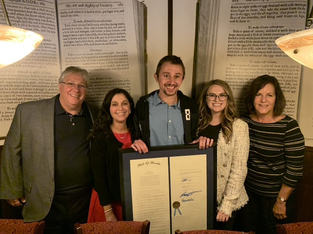 Five smiling people pose indoors with a framed document. The background features large text pages on the wall. Warm lighting.