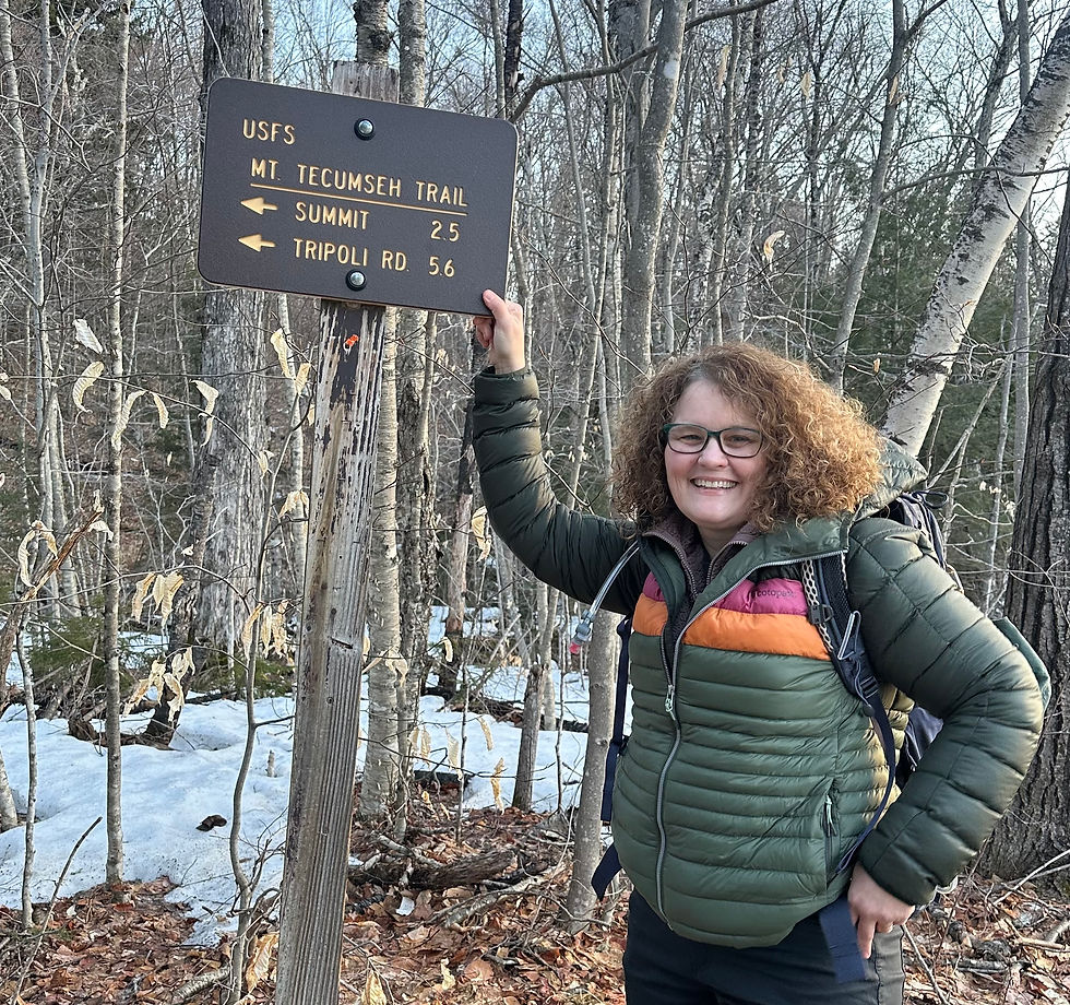 Smiling hiker in colorful jacket holds Mt. Tecumseh Trail sign in snowy forest. Trails to Summit and Tripoli Rd marked. Wooded, winter setting.