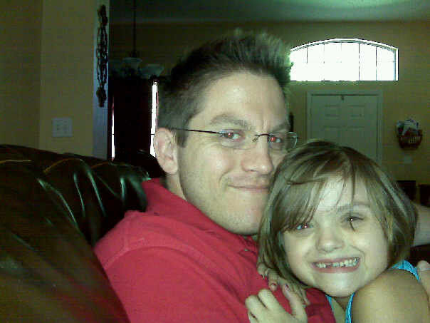 Man in red shirt and child with short hair smiling and hugging on a brown couch. A door and arched window are visible in the background.