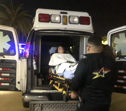 Woman on stretcher being loaded into ambulance by a paramedic at night. Palm tree in background, ambulance lights illuminate the scene.