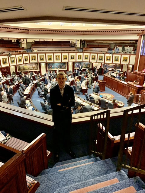 Man standing inside a large legislative chamber, filled with seated and standing people. Walls display framed pictures. Blue carpet.