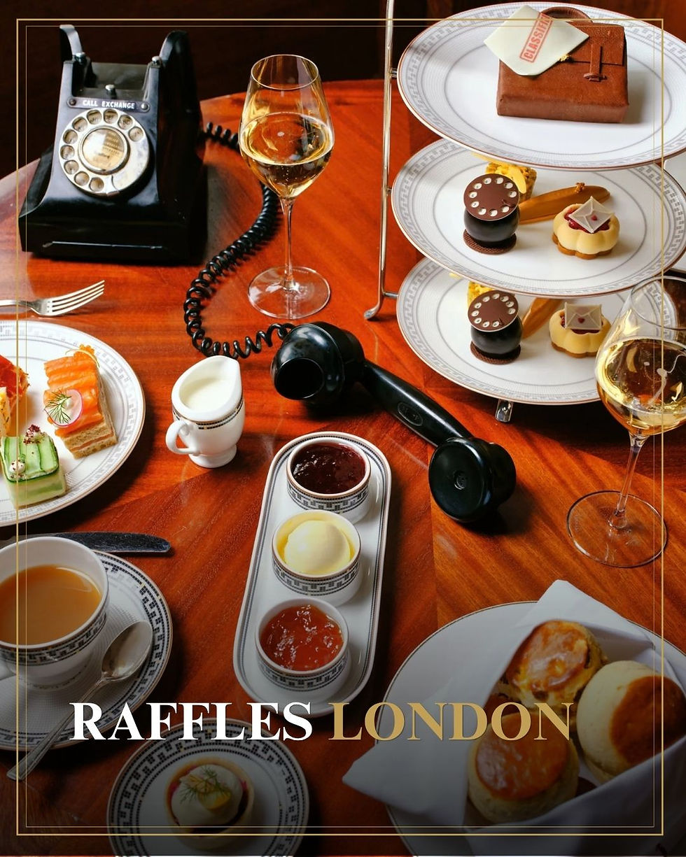 Luxe afternoon tea op houten tafel met rotarytelefoon, wijn, desserts en Raffles London-tekst.