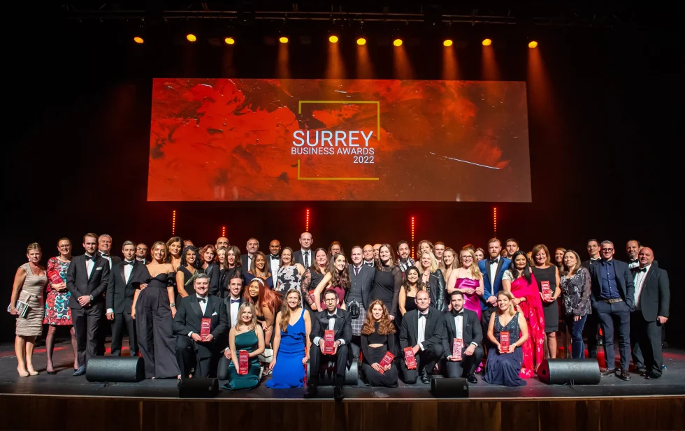 The crowd on stage. Text reads "Surrey Business Awards 2022" on screen behind them. Many smiling and looking towards the camera.