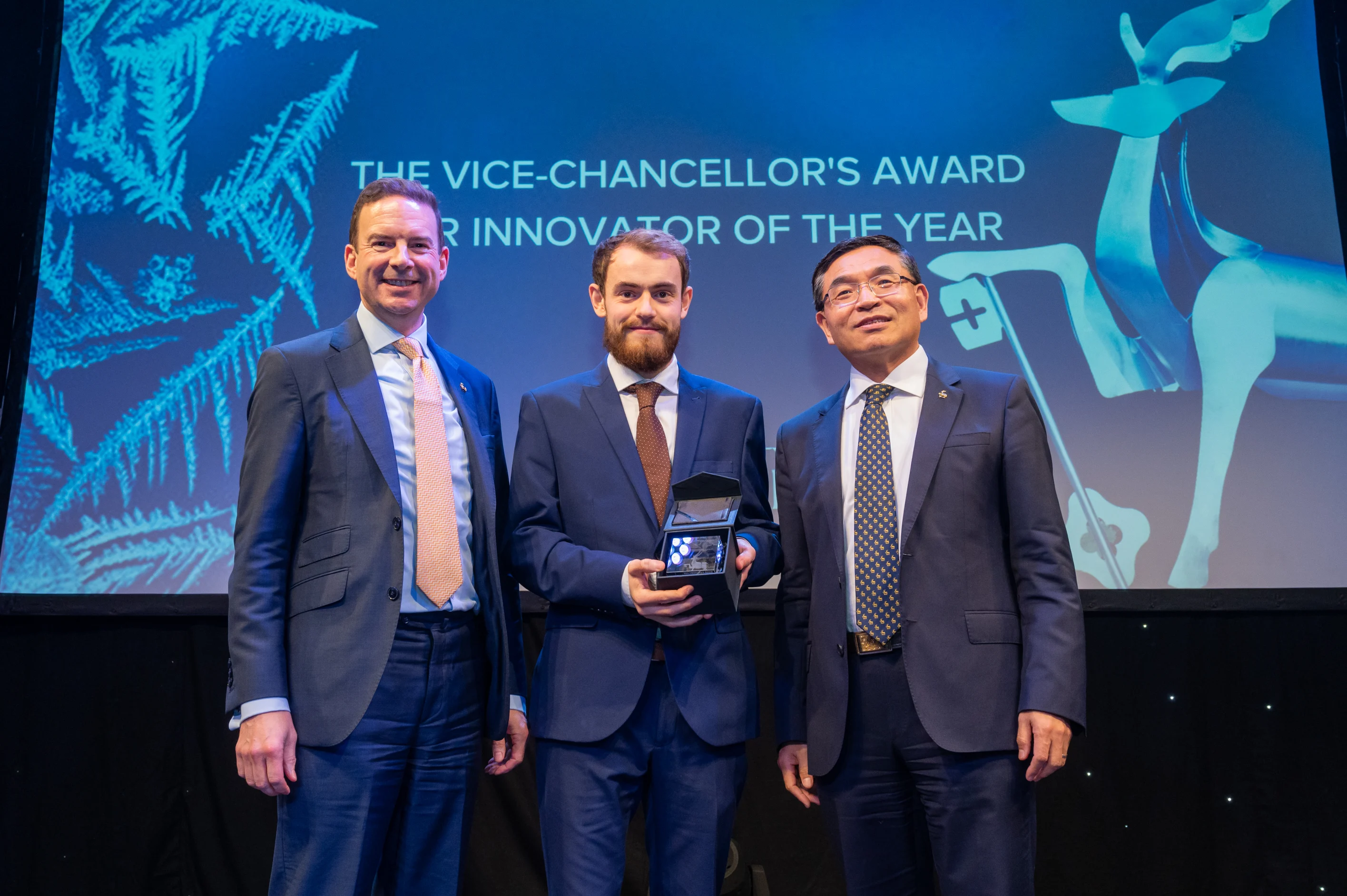 Three men standing on a stage, with the one in the middle holding an award.