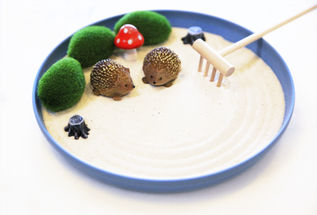 Two small hedgehog figurines on white sand in a blue tray, surrounded by green moss, a red mushroom, and a rake, creating a zen garden.