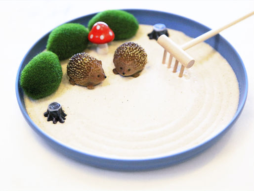 Two small hedgehog figurines on white sand in a blue tray, surrounded by green moss, a red mushroom, and a rake, creating a zen garden.