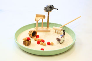 Mini cat figurines play in a sandy tray with a scratching post, rake, and colorful pom-poms. A relaxing crafting scene with soft tones.