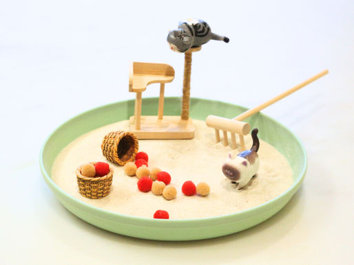 Mini cat figurines play in a sandy tray with a scratching post, rake, and colorful pom-poms. A relaxing crafting scene with soft tones.
